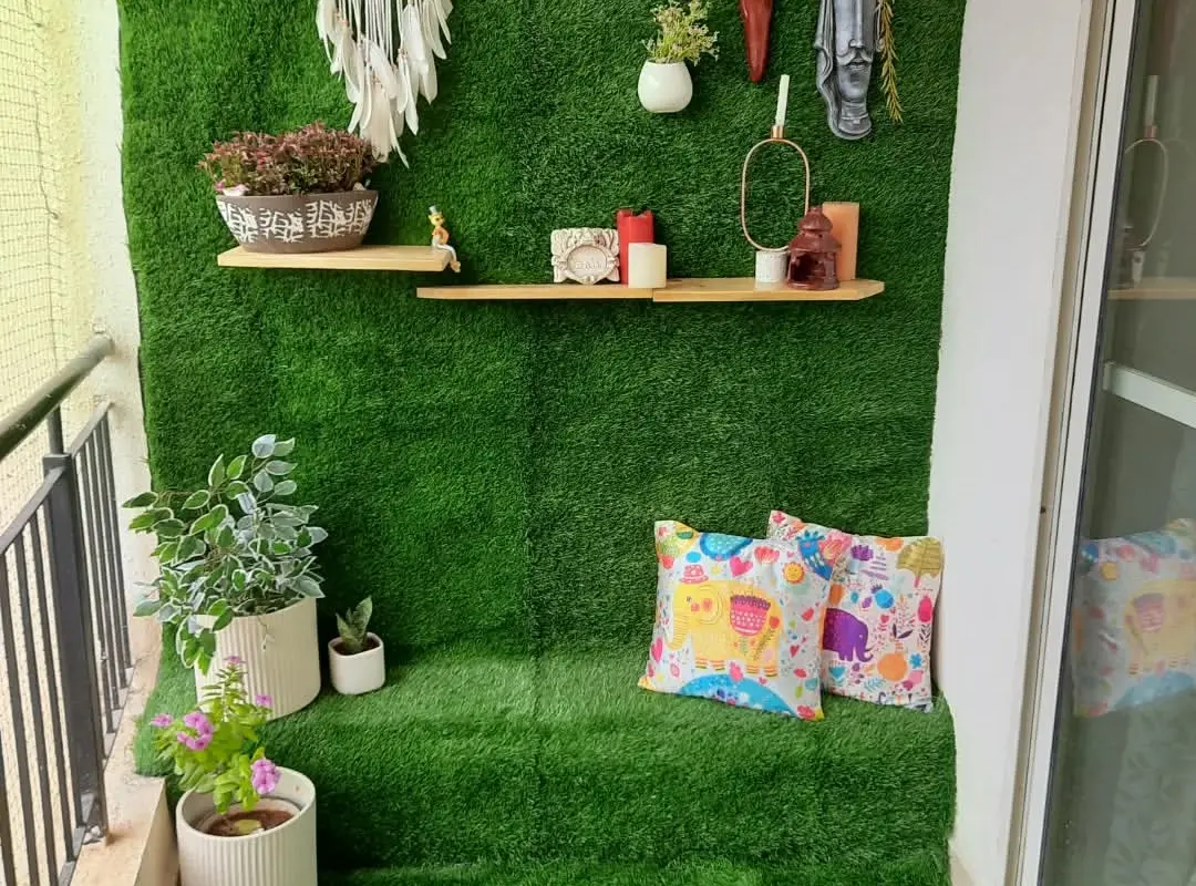 A narrow balcony with artificial grass across the floor and up the back wall, two pine shelves with plants, hanging masks and cushions