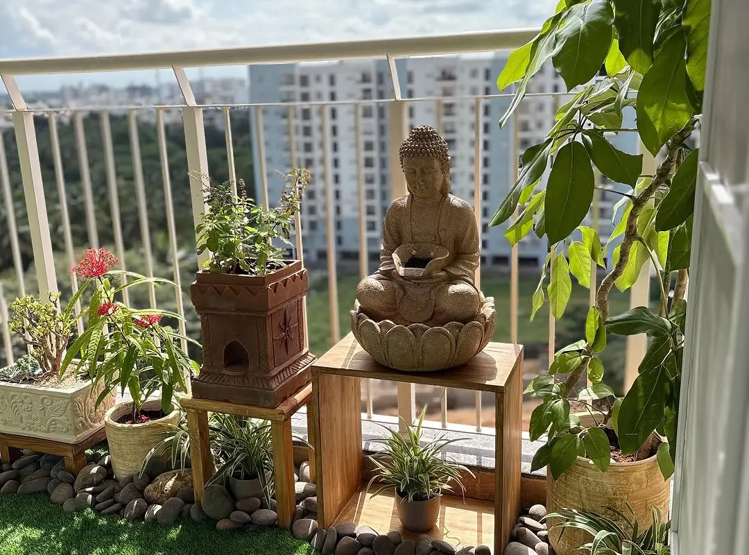 A sunny high-rise balcony with flowering and leafy plants in big terracotta and cream planters on wooden stands, a stone Buddha fountain, river pebbles and artificial grass, behind a white railing
