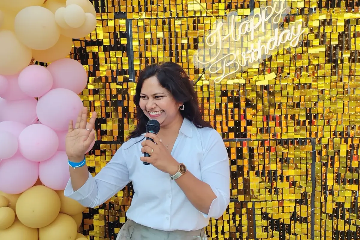 A host in a white shirt and a pale skirt laughing and waving, microphone in hand, in front of a gold sequin wall with a Happy Birthday neon sign and white, mustard and pink balloons