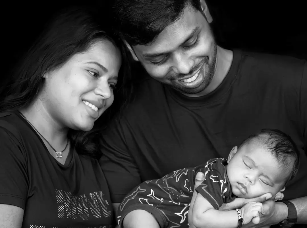 Black and white: both parents, heads together, looking down at their sleeping newborn