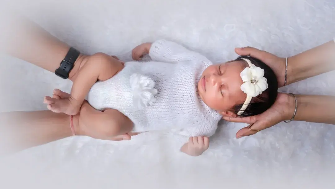 Two pairs of adult hands supporting a newborn from above, one cupping the head and one under the legs