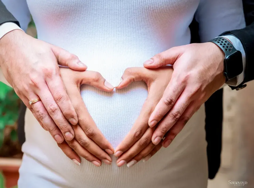 Two pairs of hands, hers and her husband's, making a heart shape on her bump, with no faces in the frame