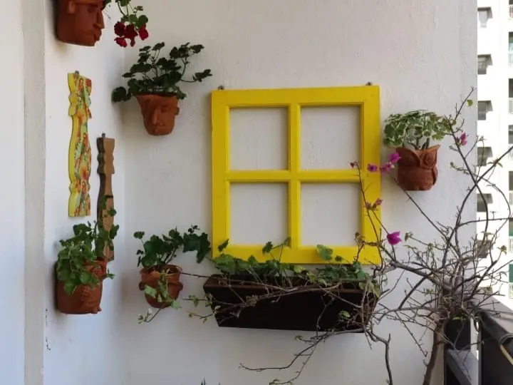 Terracotta face pots with geraniums and trailing plants fixed to a white balcony wall around a yellow window frame, with a long planter below and a bougainvillea at the edge