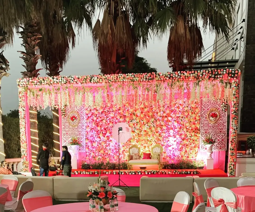 Planning a grand shadi? We design breathtaking wedding stages like this one, featuring a massive wall of pink and white flowers, elegant seating for the couple, and professional lighting to create a romantic, picture-perfect setting.