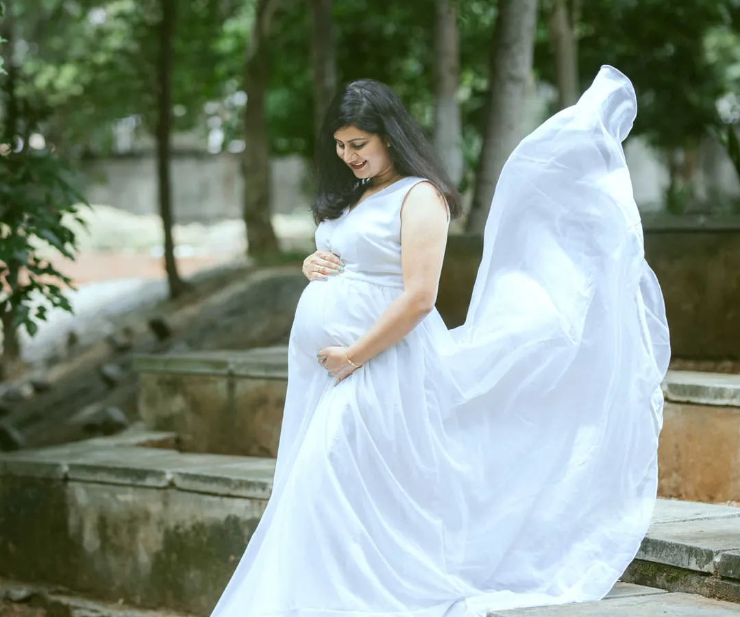 An expectant mother looking serene in a flowing white gown during an outdoor maternity session. The natural park setting and the gentle toss of the fabric create a timeless and angelic portrait.