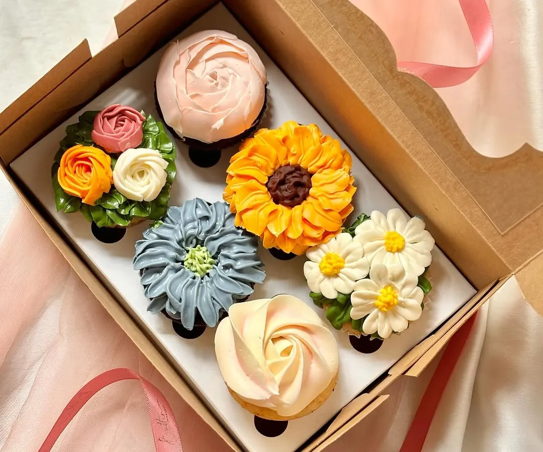 A gift box of six cupcakes decorated with intricate, multi-toned buttercream flowers. The collection displays a variety of floral piping techniques, including a sunflower, daisies, a rose, and other blossoms, perfect for elegant events like birthdays or anniversaries.