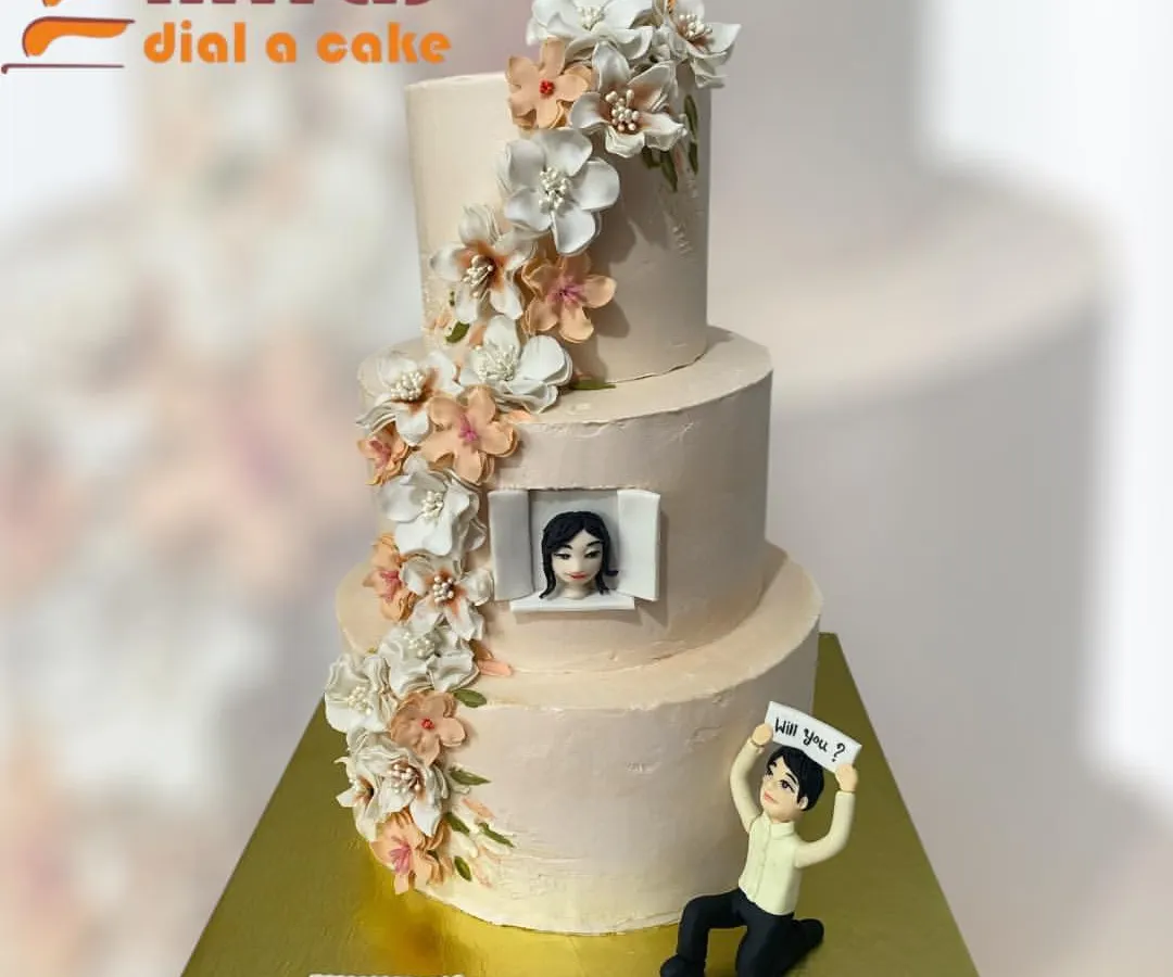 A cake that tells a love story. This elegant, three-tiered engagement cake captures a beautiful proposal moment, complete with a custom figurine, a photo of the happy couple, and a cascade of delicate, handmade sugar flowers.