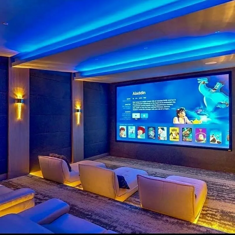 This contemporary media room is designed for both family enjoyment and critical viewing, featuring modern lounge seating with integrated floor lighting. The large-format screen is complemented by blue ambient cove lighting, creating an inviting atmosphere while ensuring excellent picture quality for everything from animated films to blockbuster movies.