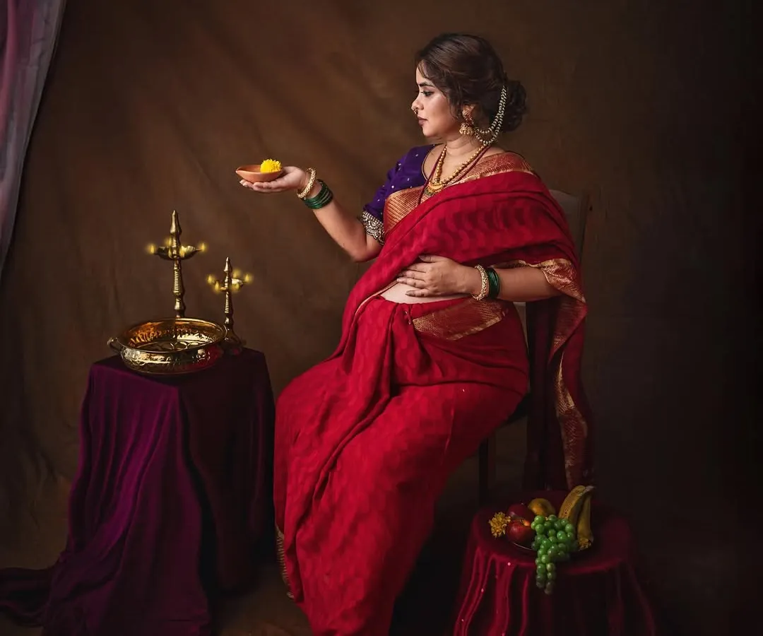 Inspired by the timeless paintings of Raja Ravi Varma, this studio portrait captures the divine grace of motherhood. Draped in a traditional red saree, this mother-to-be holds a 'laddoo', symbolizing the sweetness of new beginnings.