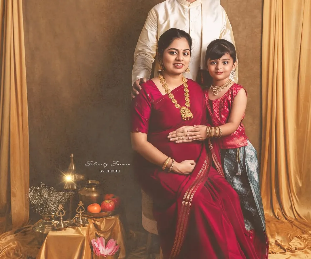 A regal family portrait celebrating a traditional Indian maternity journey. The mother, father, and daughter are dressed in elegant ethnic wear, surrounded by cultural props like brass vessels and lotuses, creating a timeless memory.