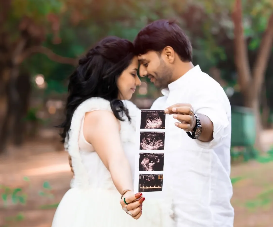 An intimate outdoor portrait of an expecting couple, touching foreheads in a moment of quiet connection. The father-to-be holds up the first glimpse of their baby: a strip of sonogram photos, symbolizing the start of their family journey.