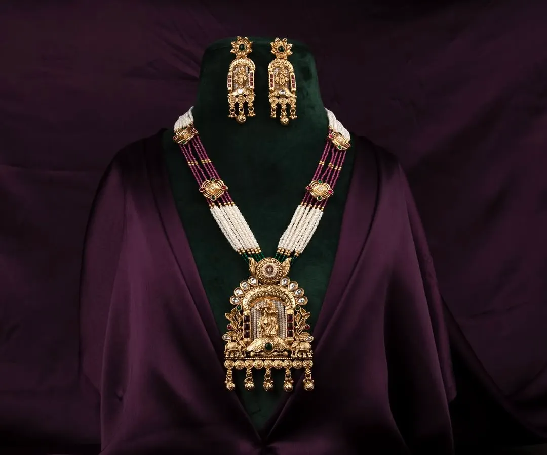 An opulent traditional Indian necklace and earring set, professionally displayed on a bust against a rich, dark purple satin background to highlight the intricate gold work and gemstones.