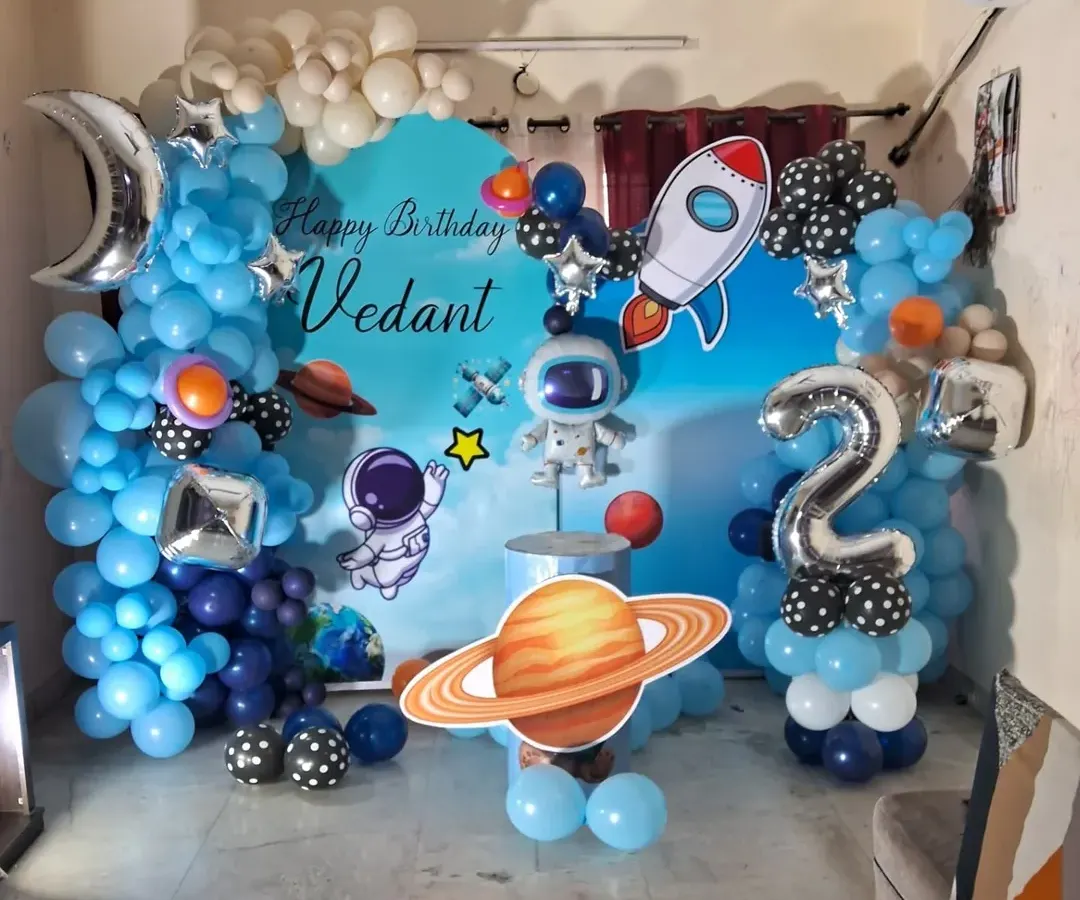 For this space-themed birthday party for Vedant, I created a custom backdrop with astronauts and planets, surrounded by a blue and silver balloon galaxy. The setup includes a large light-up marquee number '2' and a rocket ship cutout, making it an out-of-this-world experience for the kids.