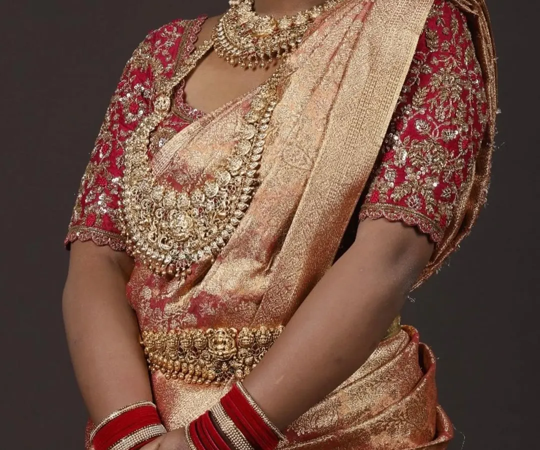 This is a classic South Indian muhurtham look. The rich red and gold Kanchipuram silk saree is paired with a custom-designed blouse featuring intricate zardozi and maggam embroidery, creating a timeless bridal outfit available for rent.