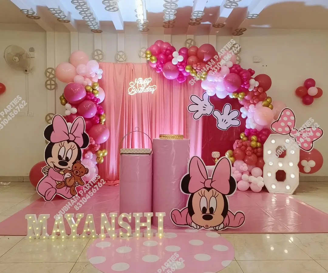 A full-themed Minnie Mouse birthday party decoration for a little one's special day. This setup includes large character cutouts, a pink fabric backdrop, a balloon arch in shades of pink and red, and marquee letters spelling out the birthday girl's name.