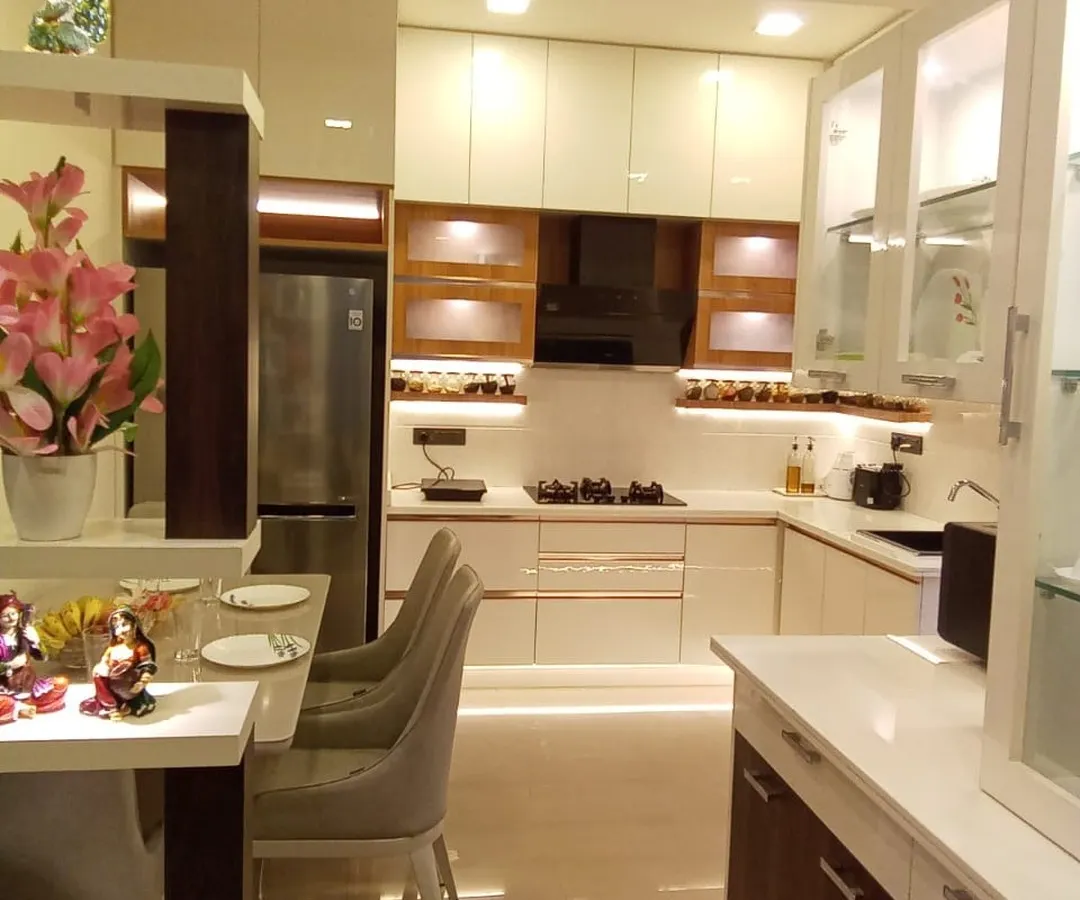 This bright and airy L-shaped modular kitchen is designed for both style and function. It features off-white and wood-finish cabinets, frosted glass shutters, and a compact breakfast counter, making it a perfect hub for the family.
