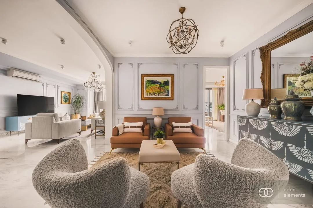 This is a perspective of a neoclassical living room, showcasing a masterful blend of classic elegance and modern comfort. The plush, textured armchairs in the foreground invite you into a space defined by symmetrical design, ornate wall moldings, and a grand, gilded mirror that amplifies the room's luxurious feel.
