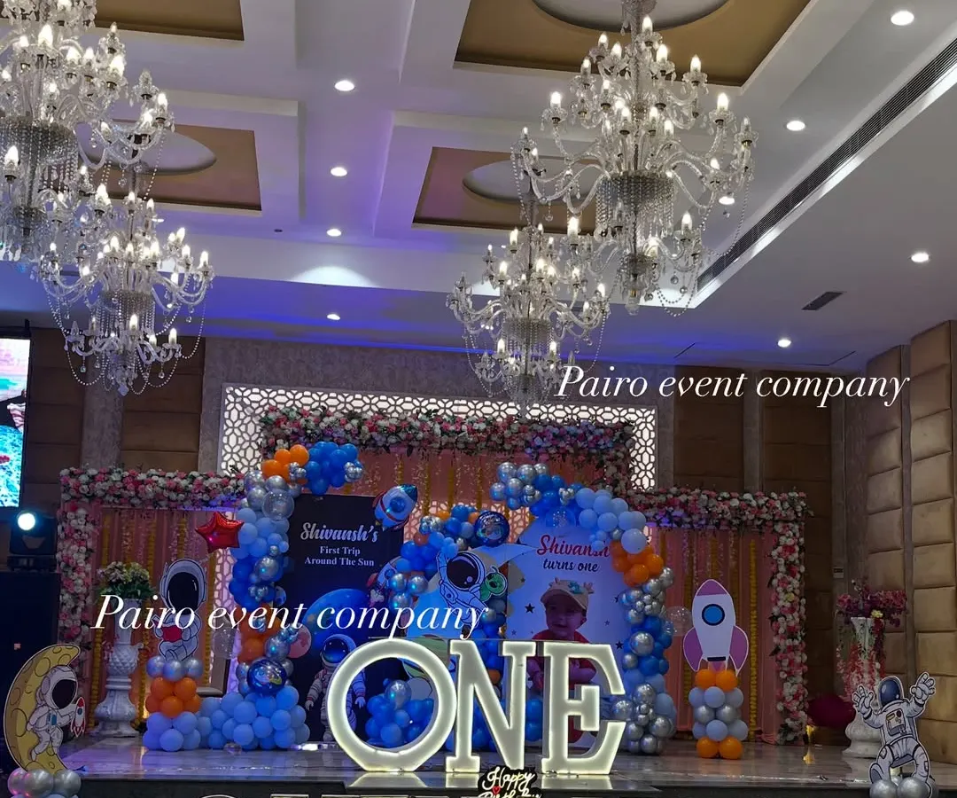 For this first birthday, we went to outer space. This wide shot shows the complete stage decoration in a banquet hall, featuring large marquee letters for the child's name and age, a custom space-themed backdrop, and extensive balloon work to create an immersive experience.