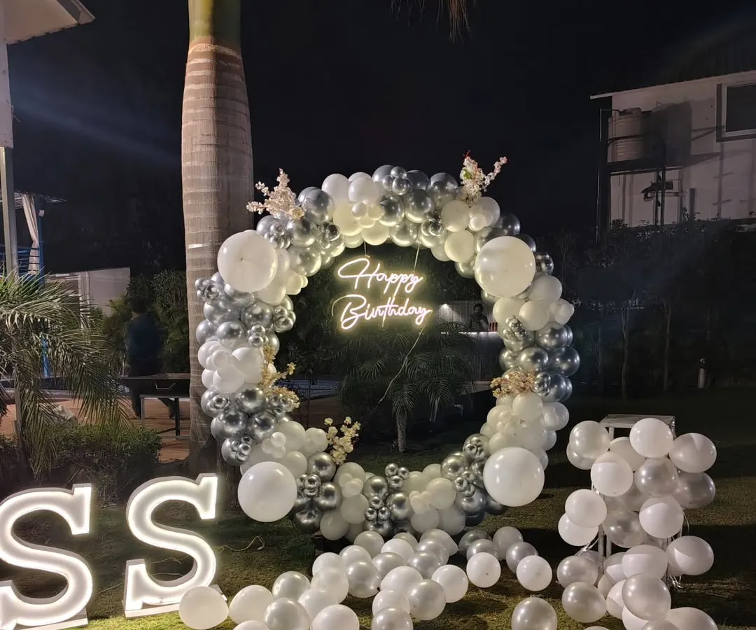 This outdoor birthday setup shows how I create a premium look for night events. The highlight is a large circular balloon garland made of silver and white chrome balloons, with a custom 'Happy Birthday' neon sign and large light-up initials for a personal touch.