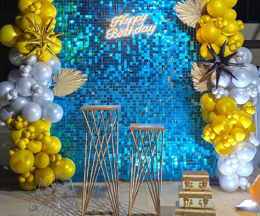 For a touch of glamour, we designed this stunning birthday backdrop. It features a vibrant blue shimmer wall that catches the light beautifully, framed by bold yellow and silver balloon garlands and a bright "Happy Birthday" neon sign.