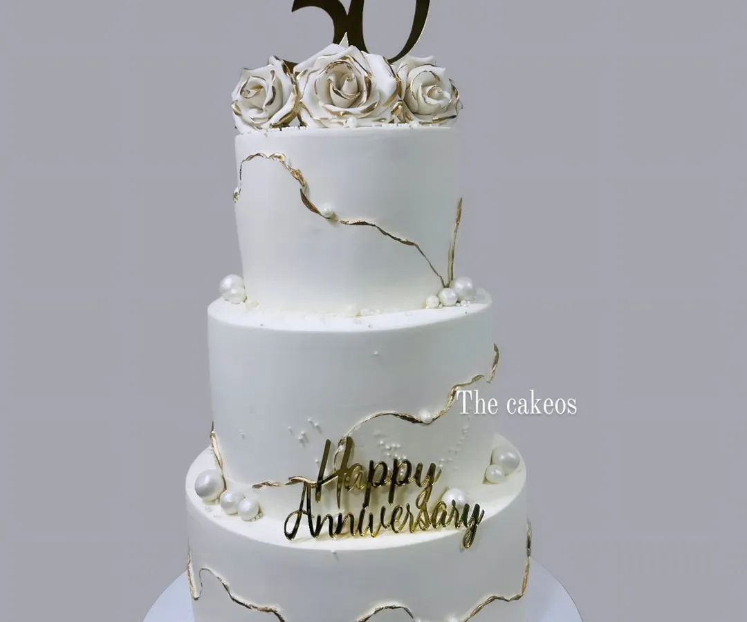 A stunning three-tier eggless cake created to celebrate a golden 50th anniversary. The design features elegant white fondant with delicate gold kintsugi-style cracks, pearls, and handcrafted sugar roses, perfect for a landmark celebration.