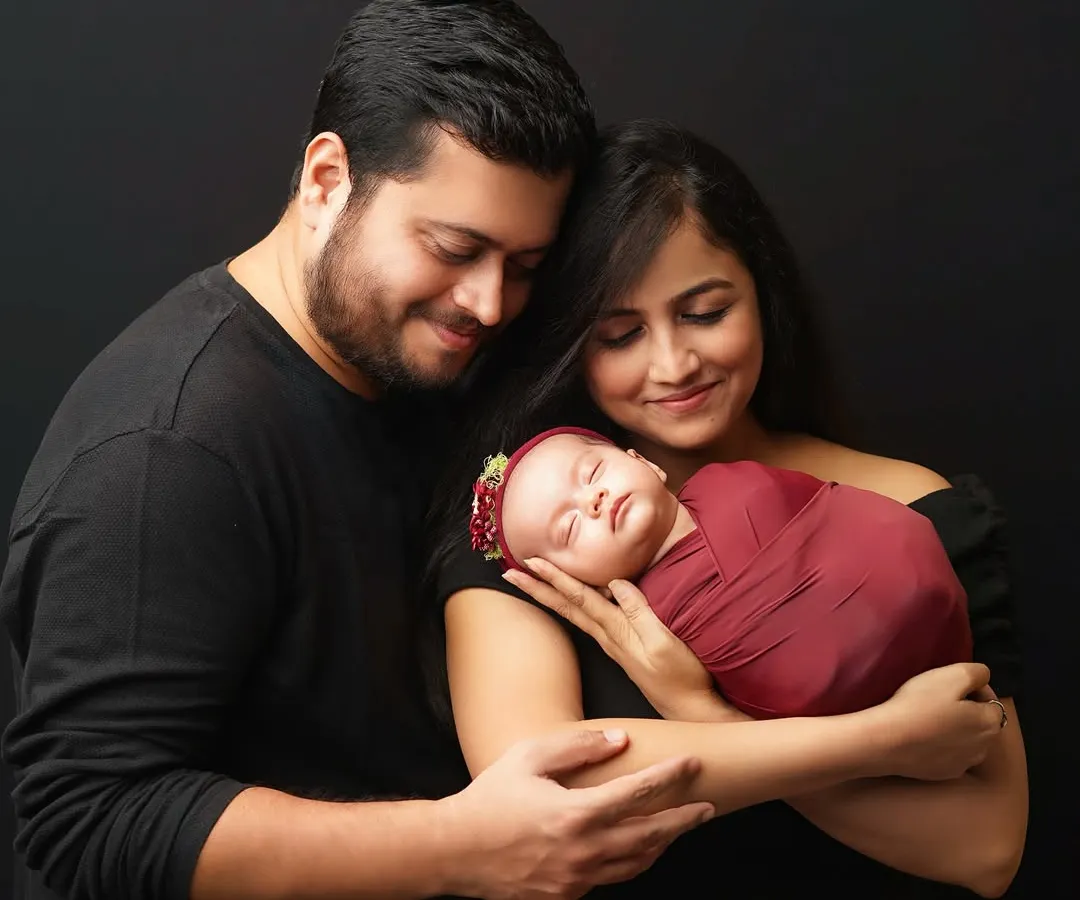 A classic family portrait against a simple black background. The new parents are smiling as they gaze down at their sleeping newborn, who is swaddled in a rich maroon wrap with a matching floral headband.