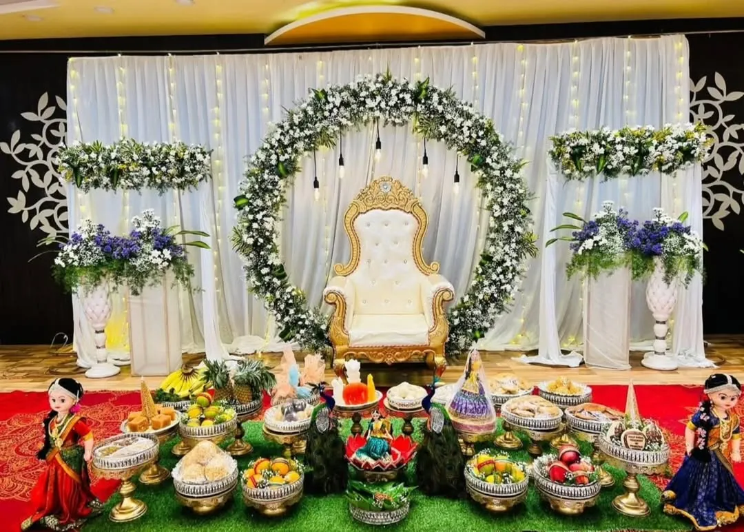 For a more modern and elegant look, I designed this Half Saree function stage with a beautiful white floral arch, hanging lights, and coordinated flower stands. The seer plates are arranged on a lush green mat, blending tradition with contemporary style.