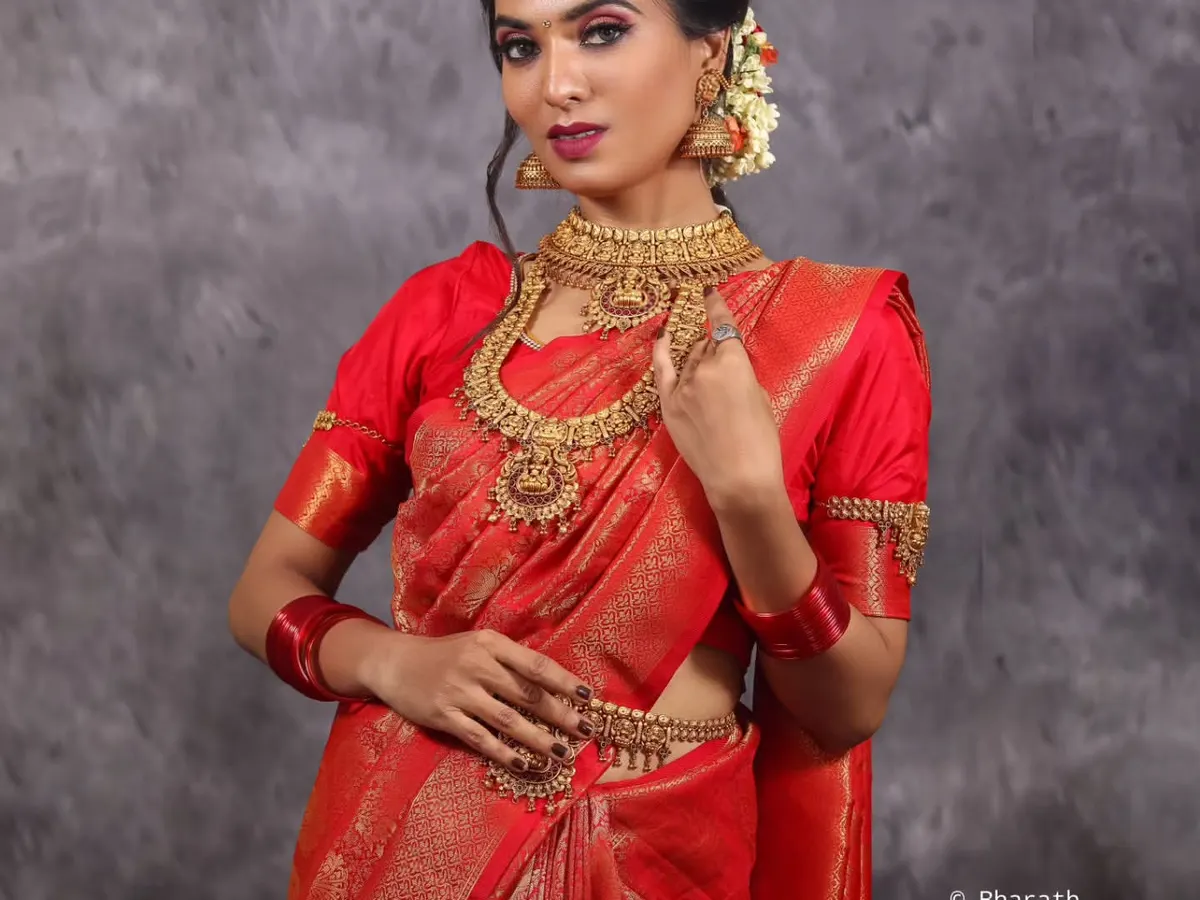 A classic studio portrait showcasing a traditional bridal look. The controlled lighting highlights the rich color of the red saree and the intricate details of the temple jewelry.