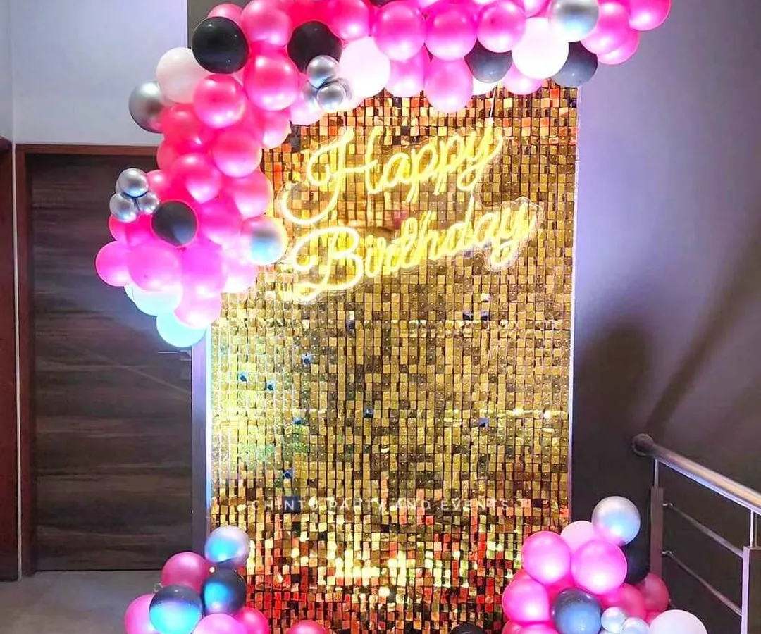 A modern and glamorous birthday photo booth with a gold shimmer sequin wall. The setup is completed with a custom neon sign and a vibrant pink and black balloon arch, perfect for chic celebrations.