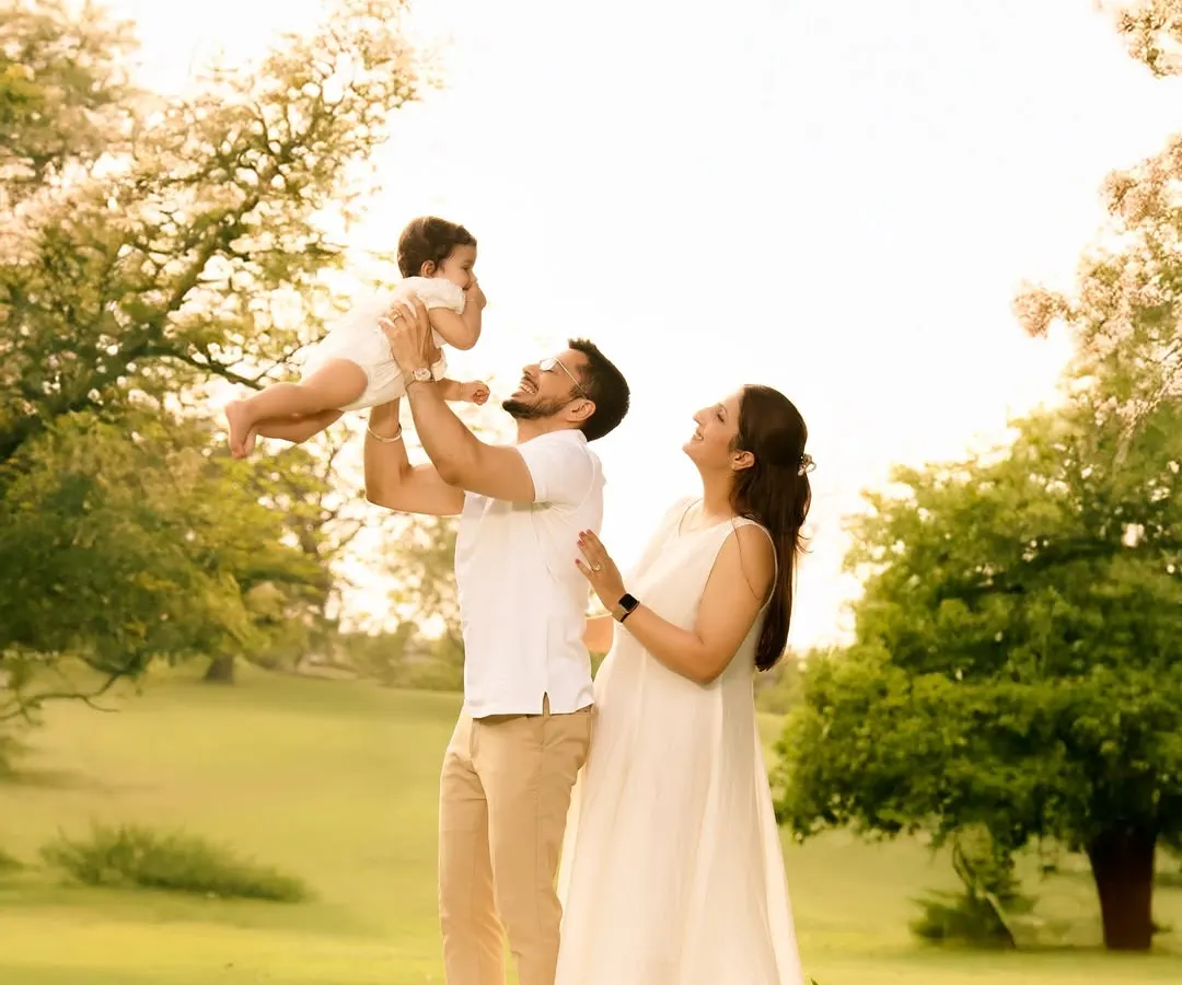 A beautiful, sunlit moment of a family enjoying a picnic in a lush green park. The father joyfully lifts his baby into the air as the mother looks on with a smile, perfectly capturing the warmth and happiness of candid family life.