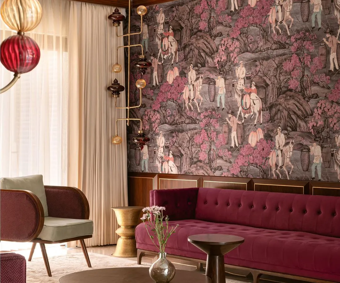 We believe in fearless design, as shown in this living room from our 'Tinted Tapestries' project. A rich plum velvet sofa is set against a dramatic, hand-painted wallpaper, balanced by sculptural lighting and tables to create a space that is both maximalist and meticulously curated.