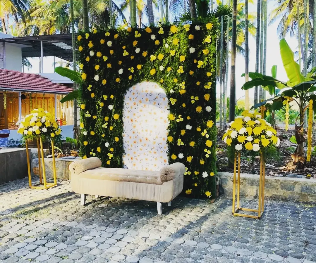 For this outdoor haldi ceremony, I created a vibrant photo spot with a lush greenery wall dotted with yellow and white flowers. The design includes a comfortable seating area and matching floral stands, all set against a beautiful natural backdrop of palm trees.