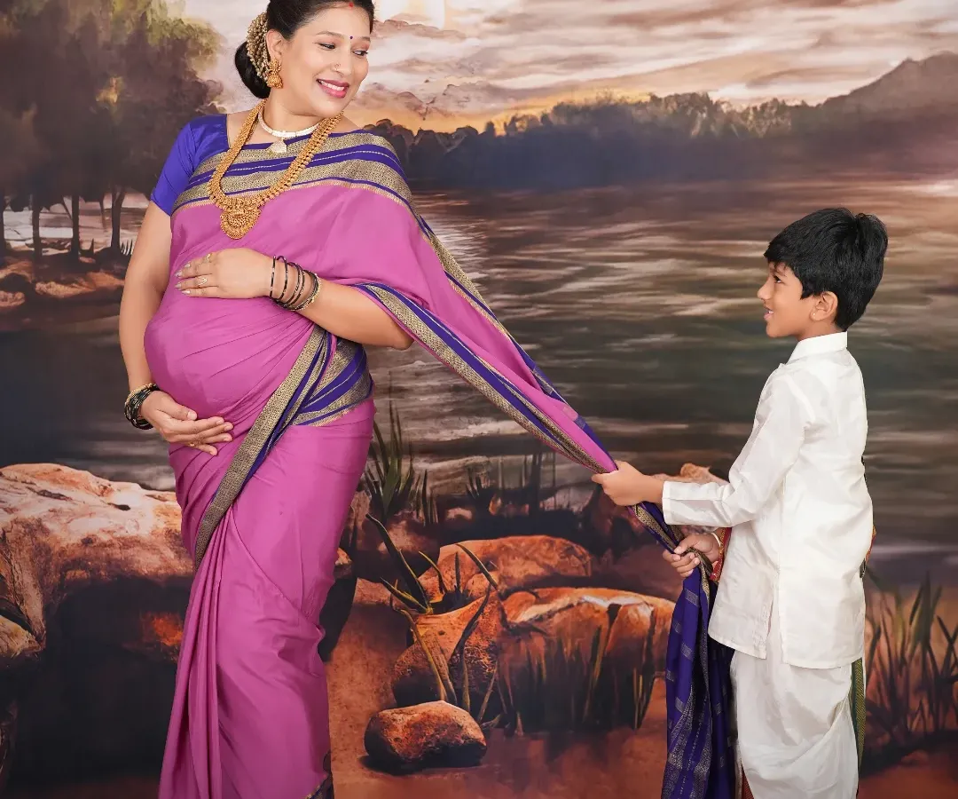 This portrait is inspired by the timeless art of Raja Ravi Varma. The mother-to-be, dressed in a traditional pink and blue saree, shares a gentle moment with her son against a classic, painted backdrop, creating a piece of art that feels both regal and personal.