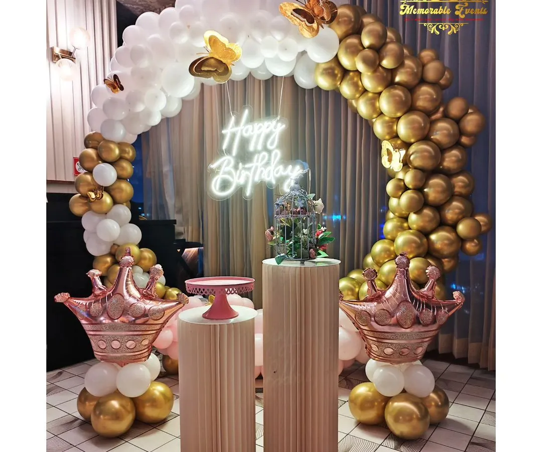 This elegant birthday decoration features a beautiful arch of white and chrome gold balloons, accented with butterfly details and rose gold crown balloons. The setup is completed with stylish plinths and a modern neon 'Happy Birthday' sign.