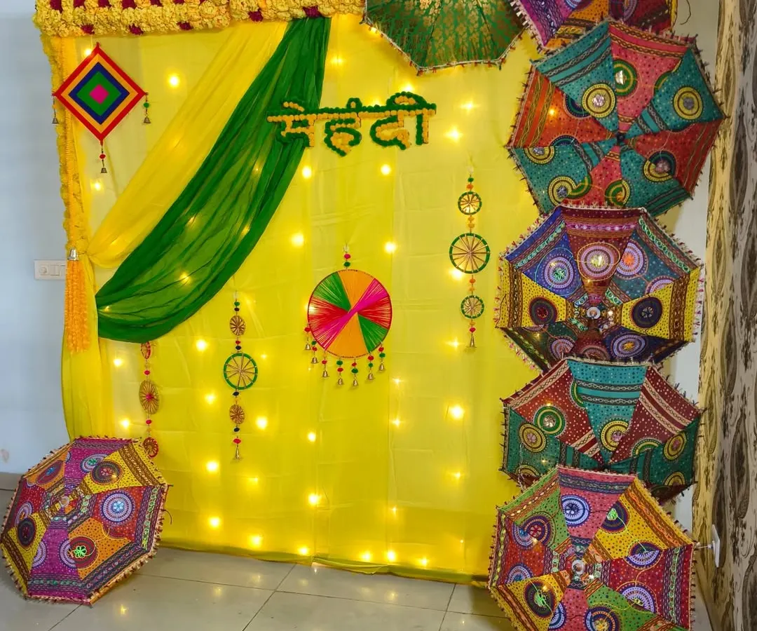 For a traditional Mehendi ceremony, I create a vibrant backdrop using yellow and green fabric drapes, warm fairy lights, and a collection of colorful Rajasthani umbrellas with mirror work. The Hindi "Mehendi" lettering adds an authentic touch.