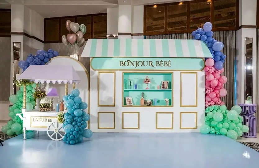 An elegant Parisian-themed baby shower setup, showcasing a custom "Bonjour Bébé" patisserie-style backdrop in mint green and white with gold accents. The display includes a charming Ladurée-inspired cart and is flanked by colorful balloon clusters in blue, pink, and green, creating a chic and welcoming atmosphere.