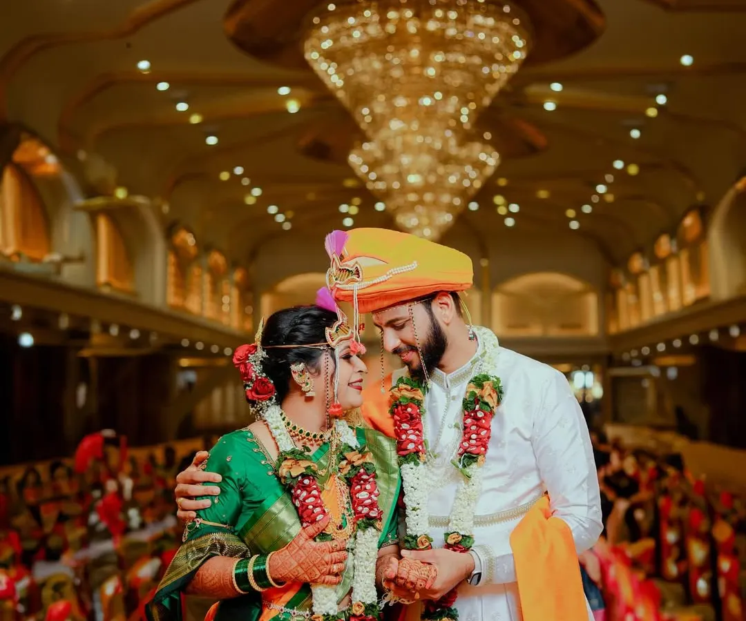 Capturing the grandeur of a traditional Maharashtrian wedding. The couple shares a look under a magnificent chandelier, surrounded by the warmth and opulence of their celebration, a perfect blend of tradition and splendor.