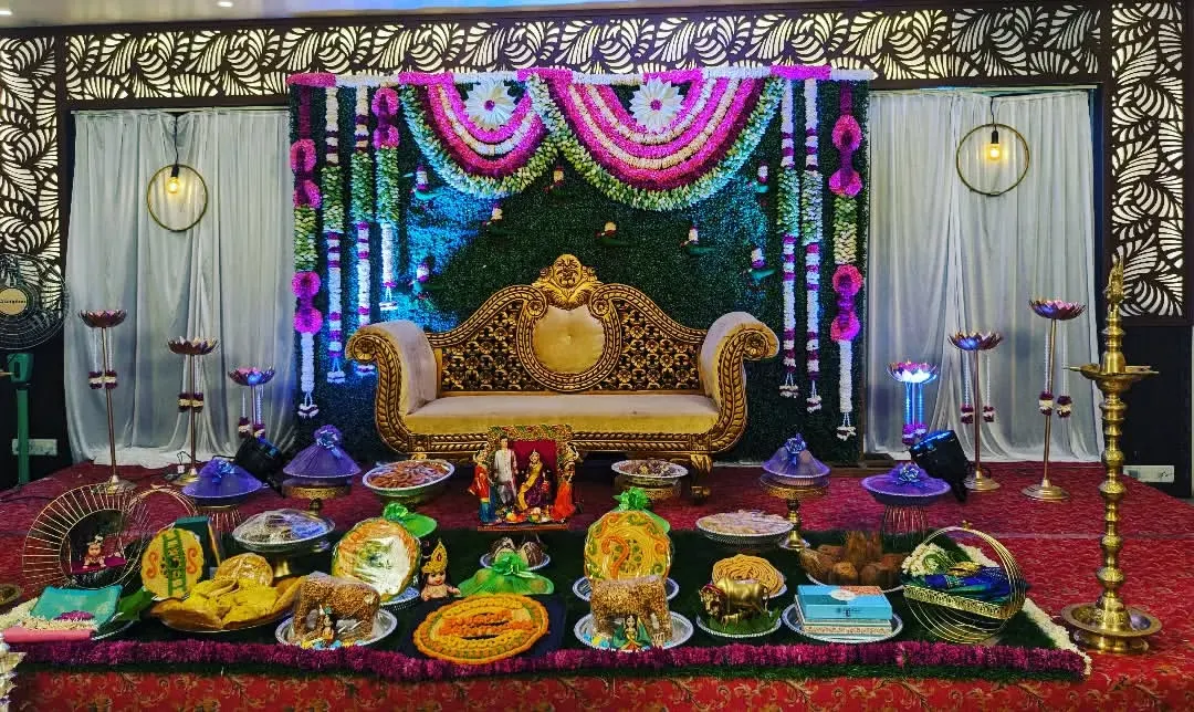 A grand and traditional Seemantham ceremony stage. We designed this with a lush green and floral backdrop, an ornate golden sofa, and a full display of beautifully arranged ceremonial plates.
