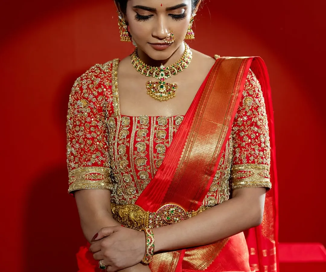 A close-up of a classic South Indian bridal look, featuring a vibrant red Kanjeevaram saree paired with a heavily embroidered blouse. The rich gold zari work and traditional motifs on the blouse showcase the pinnacle of heritage craftsmanship, perfect for a bride seeking authentic grandeur.