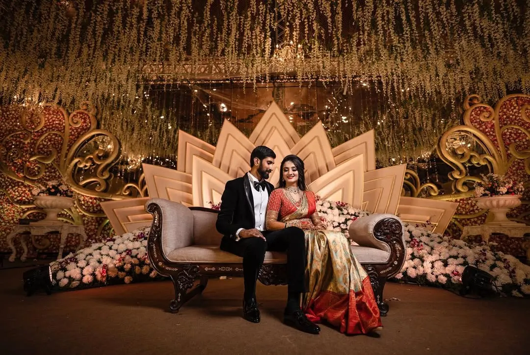 A formally dressed couple is elegantly posed on a luxurious chaise lounge against a grand, lotus-themed stage at their wedding reception. We specialize in capturing the scale and design of large-scale wedding events.