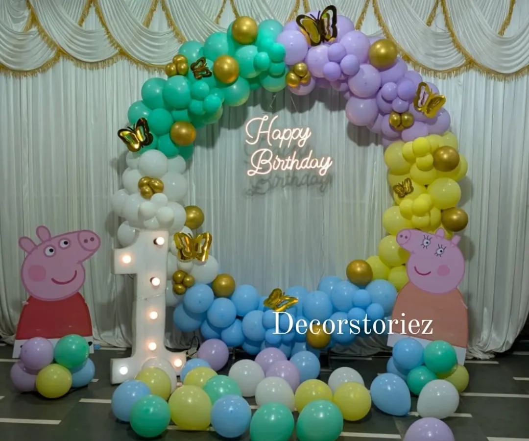 A complete Peppa Pig party setup perfect for a first birthday. This features a full balloon ring in pastel colors, life-sized character cutouts, a light-up marquee number '1', and a custom 'Happy Birthday' neon sign for that extra glow.