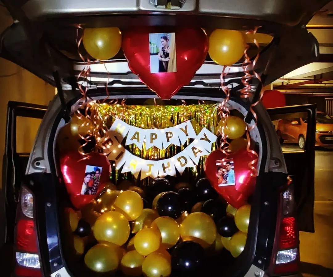 A unique car trunk birthday surprise, perfect for creating a memorable moment. This setup uses gold, black, and red balloons, a "Happy Birthday" banner, and personalized photos attached to heart-shaped foil balloons.