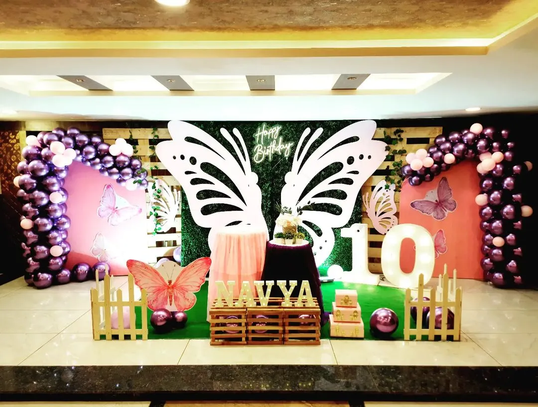 This butterfly-themed stage for Navya's 10th birthday features a lush greenery wall as the base. I added dramatic, oversized white butterfly wings as the central photo-op element, framed by balloon columns in shades of purple and pink for a vibrant, garden-like feel.