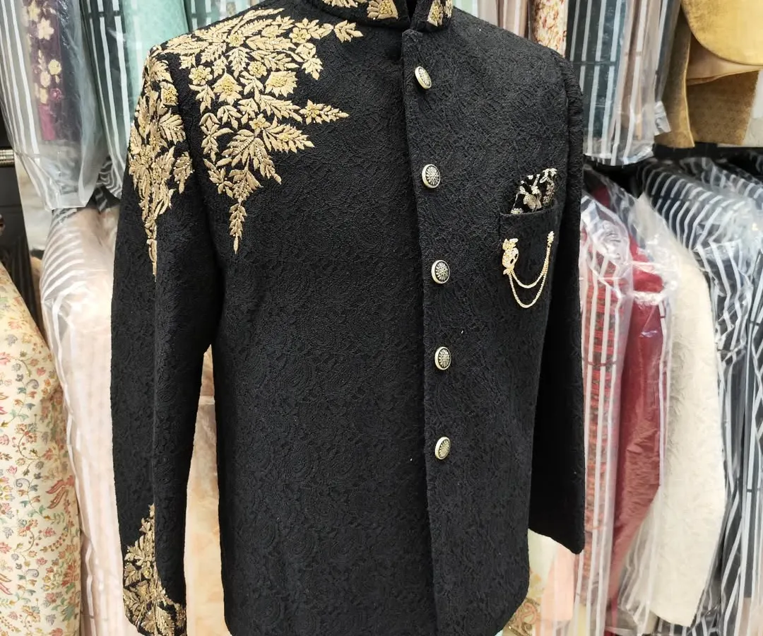 A showcase of regal craftsmanship. This black Jodhpuri suit features intricate gold hand embroidery on the shoulder and sleeve, creating a look of timeless elegance perfect for a groom or a grand event.