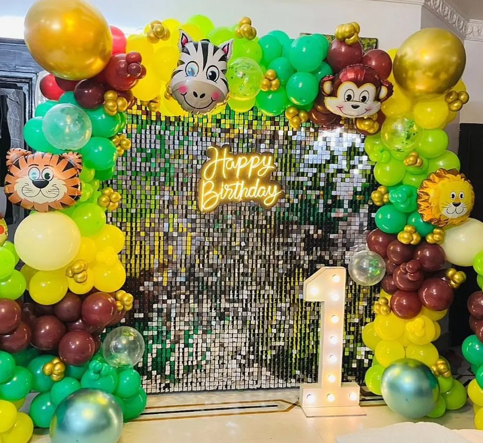 A vibrant jungle safari theme, perfect for a first birthday party. This setup features a shimmering sequin wall, a neon "Happy Birthday" sign, animal-themed foil balloons, and a large light-up marquee number '1'.