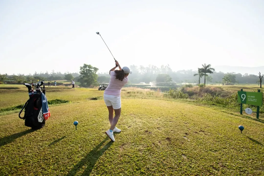 We welcome golfers of all levels and backgrounds. Here, a player is captured mid-swing, teeing off on a beautiful morning with the course's water features and lush landscape in the background.