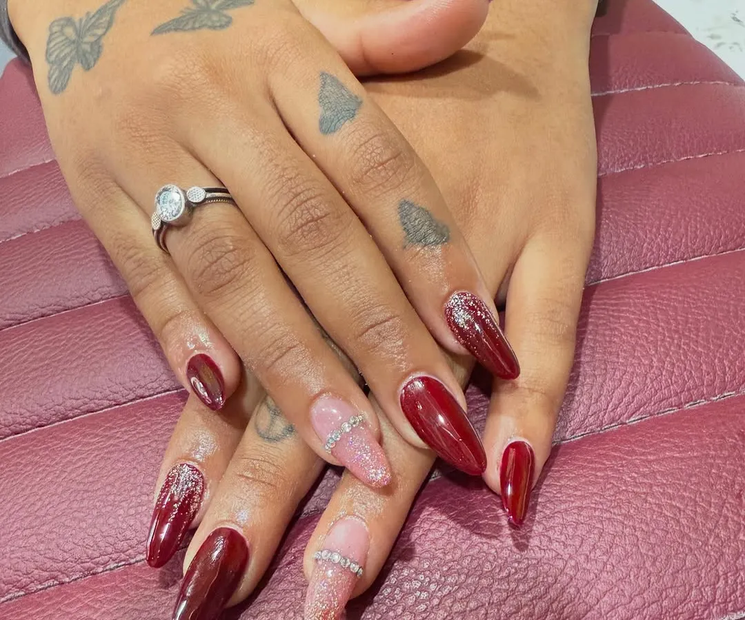 A classic look with a touch of glamour. This set features long, almond-shaped nail extensions in a deep, glossy red, with accent nails in soft pink glitter and a delicate rhinestone band for extra sparkle. The client's hand tattoos show how well body art and nail art can complement each other.