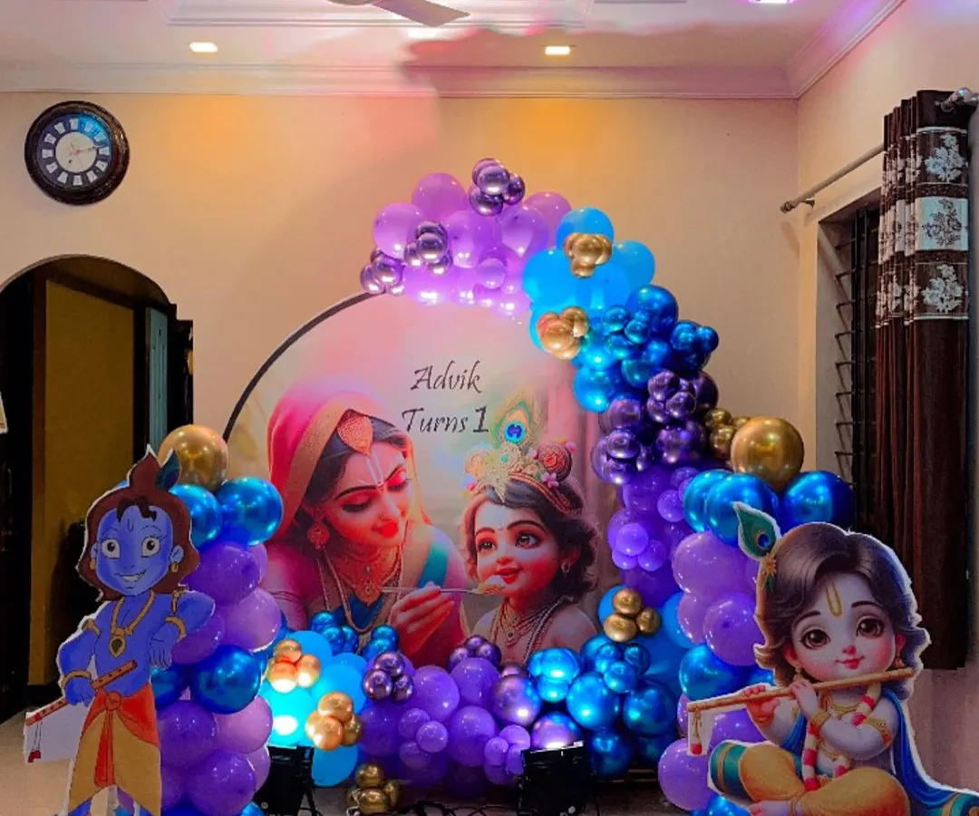This Krishna theme decoration for a first birthday creates a vibrant and divine atmosphere. The setup includes a large circular backdrop with Yashoda and Krishna, flanked by character cutouts and a rich balloon arch in shades of blue, purple, and gold.