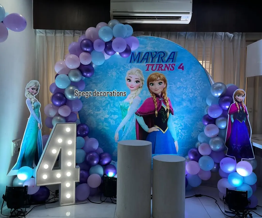 A magical Frozen-themed birthday party for a 4-year-old. This setup includes a custom circular backdrop with Anna and Elsa, life-sized character cutouts, a light-up marquee number, and a balloon arch in a wintery color palette.