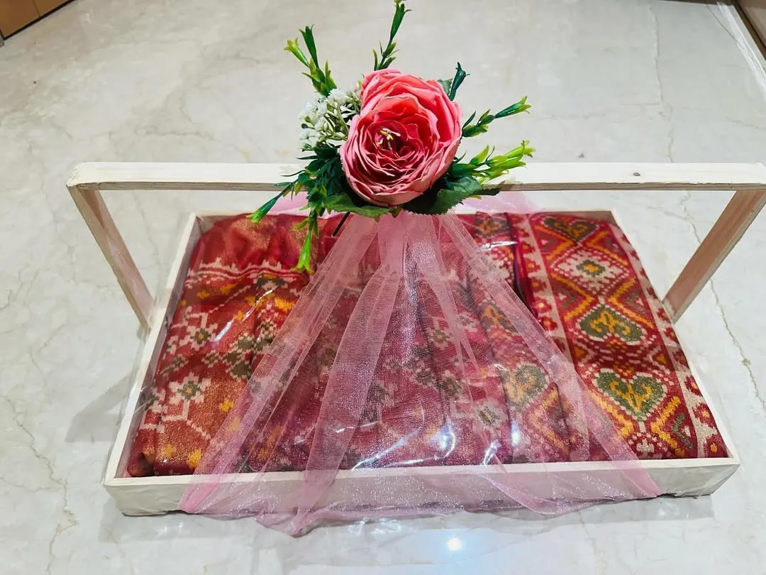 This is how I deliver a complete gift experience. A traditional Patola-style saree is carefully packed in a wooden tray with a clear acrylic top, wrapped in soft pink tulle, and finished with a floral arrangement, ready for any shagan ceremony.